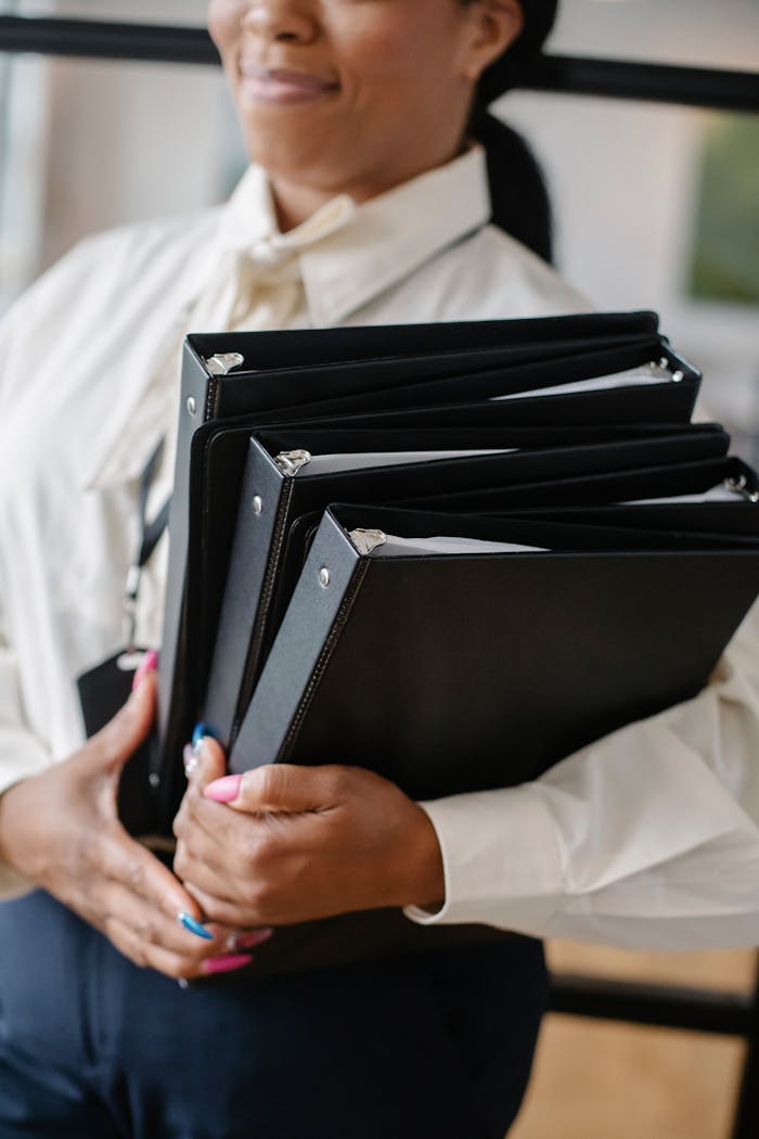Professional woman in formal attire carrying multiple black folders indoors.
